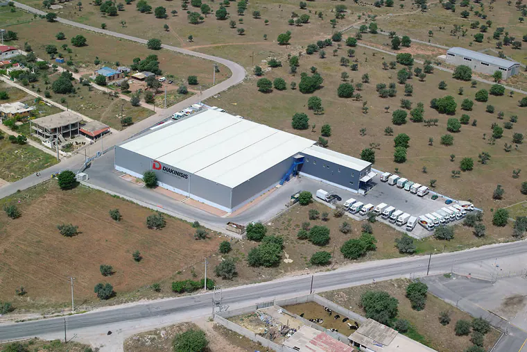 Aerial view of a logistics warehouse with a white roof and parked trucks, among scattered trees