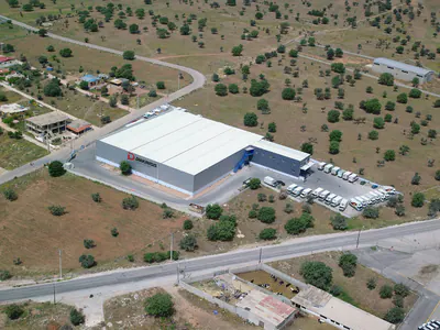Aerial view of a logistics warehouse with a white roof and parked trucks, among scattered trees