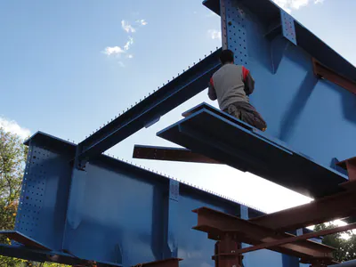 Worker kneeling on a steel beam between blue bridge girders, seen from below