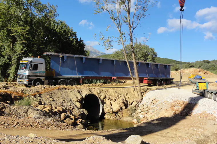 Lorry carrying a blue bridge girder past a stream culvert