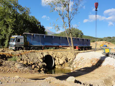 Lorry carrying a blue bridge girder past a stream culvert