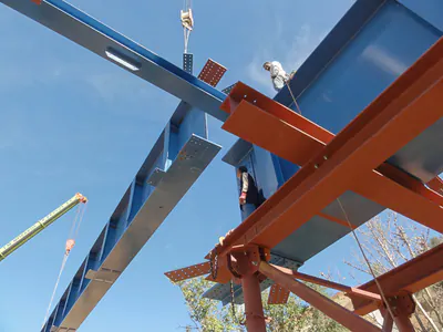 Blue bridge girders on red temporary supports, seen from below, with a crane and workers