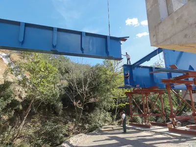 Crane lifting a blue girder over a wooded ravine, beside girders on red temporary supports