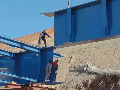 Workers beside a blue bridge girder during assembly
