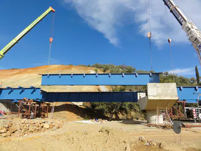 Two mobile cranes lifting a blue bridge section, with a concrete pier and temporary supports