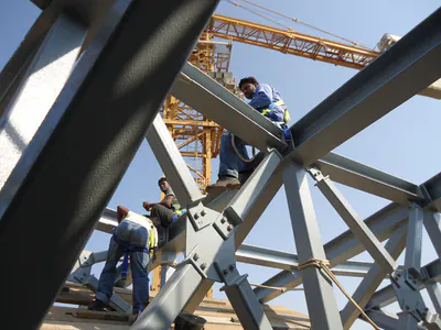 Workers on a grey steel frame with bolted joints, a tower crane behind