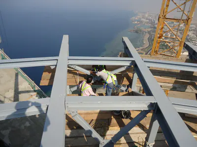 Workers on a grey steel frame, with a tower crane and the sea behind
