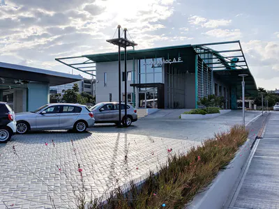 Building with teal steel canopies beside a glazed pavilion and a paved car park