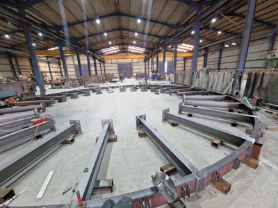 Circular steel frame laid out on the floor of a production hall, radial beams running in from a curved edge girder