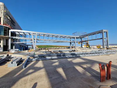 Grey steel frames of two elevated walkways reaching out from a building under construction over paved ground, steel members laid out in front
