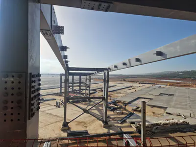 Bolted steel column and beams framing a view over a wide paved area, a braced steel frame standing below