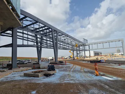 Grey steel frame of an elevated walkway seen from below beside a building, lifts at work beyond, under a cloudy sky