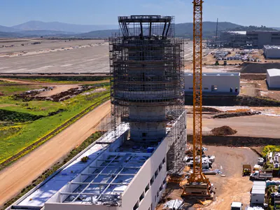 Aerial view of a round tower in scaffolding rising from a white building, a tower crane beside it and an airfield beyond
