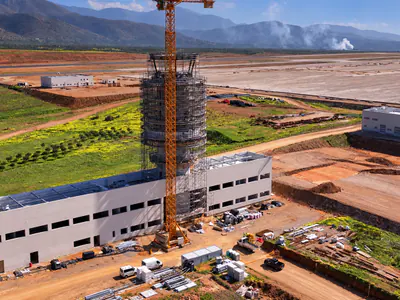 Aerial view of a tower in scaffolding rising from a long white building, a tower crane beside it, fields and mountains behind