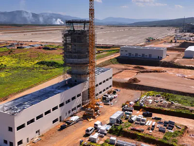 Aerial view of a tower in scaffolding above a white building, a tower crane beside it, low white buildings and an airfield behind