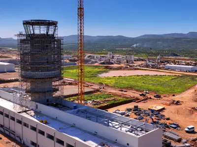 Aerial view of a tower with a glazed cab in scaffolding beside a long white building, a construction site and mountains beyond