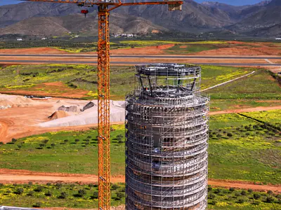 Top of a tower in scaffolding beside a tower crane, with fields, an airfield and mountains behind