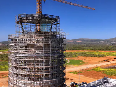Top of a round tower wrapped in scaffolding, a glazed cab at the summit, fields and hills behind