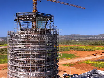 Scaffolded top of a round tower under a tower crane's jib, fields and hills beyond