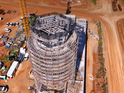 Overhead view of a round tower's open roof frame inside scaffolding, a tower crane and parked vans beside it