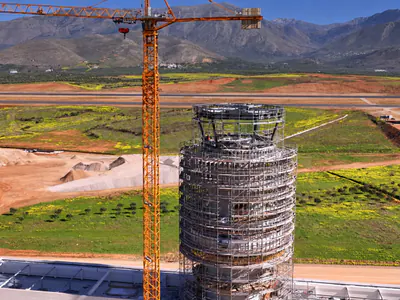 Top of a tower in scaffolding behind a white roof, a tower crane rising beside it, an airfield and mountains beyond