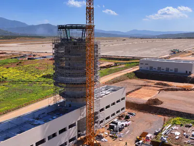 Aerial view of a tower in scaffolding above a white building, a tower crane beside it and an airfield under a cloudy sky