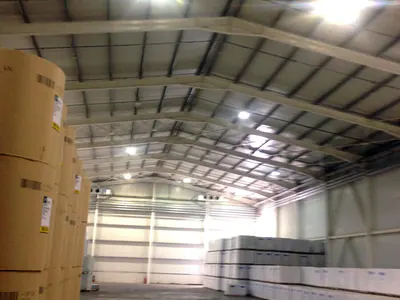 Warehouse interior with a steel roof frame, bright lights, stacked rolls and pallets