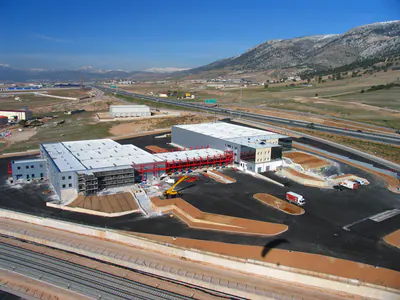 Warehouse and office buildings with a long red steel frame between them, scaffolding and a mobile crane, aerial view