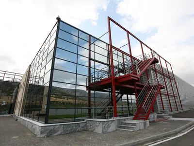 Glass-walled block with an external red steel staircase in a red steel frame, beside a metal-clad warehouse