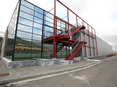 Glass-walled block with an external red steel staircase in a red steel frame, at the corner of a long warehouse