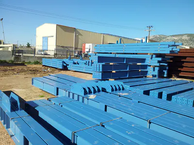 Blue-painted steel beams stacked in a yard beside a warehouse