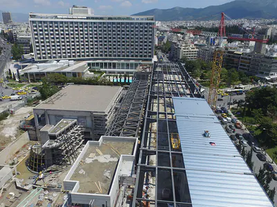 Aerial view of a building under construction with steel roof framing, among city blocks