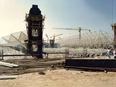 Steel space-frame roof under erection beside a tall tower wrapped in scaffolding