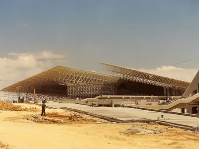 Large steel space-frame roofs seen across a sandy construction site