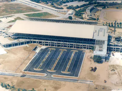 Aerial view of a large building with a partly unclad steel space-frame roof and an empty car park in front