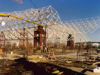 Tall towers and workers under a steel space-frame roof, with a concrete pump boom and cranes