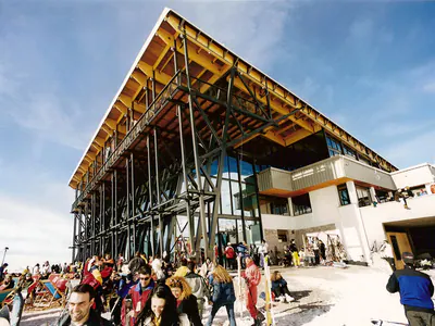 Ski centre building with a large pitched timber roof on steel columns, crowds in the snow