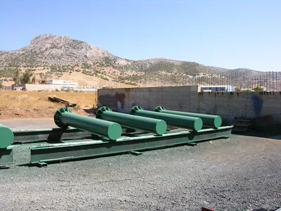 Green-painted tubular steel columns laid out in a yard, with hills behind