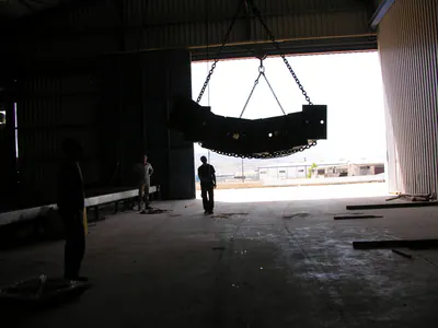 Curved steel member hanging from lifting chains inside a hall, workers in silhouette