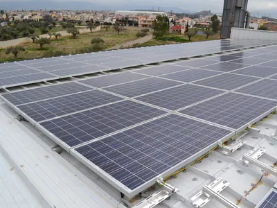Σειρές φωτοβολταϊκών πάνελ σε στέγη, με πόλη και λόφους στο βάθος